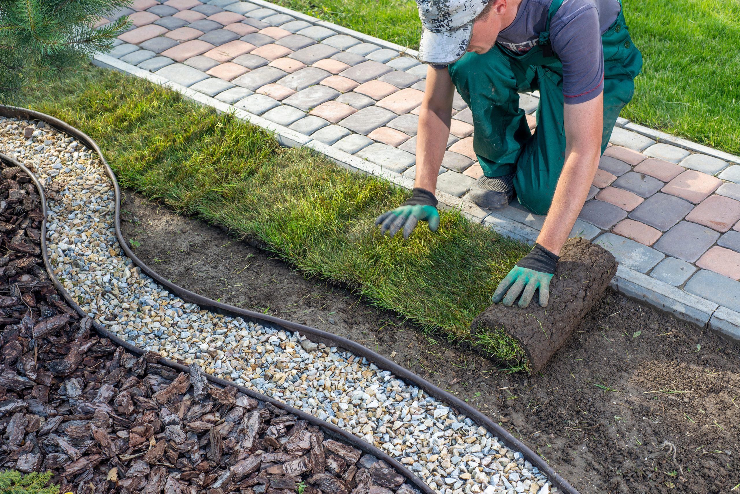 Person laying sod on the ground next to a paved path and decorative gravel border.