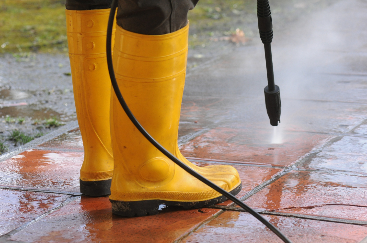 Person wearing yellow boots power washing wet red tiles outdoors.